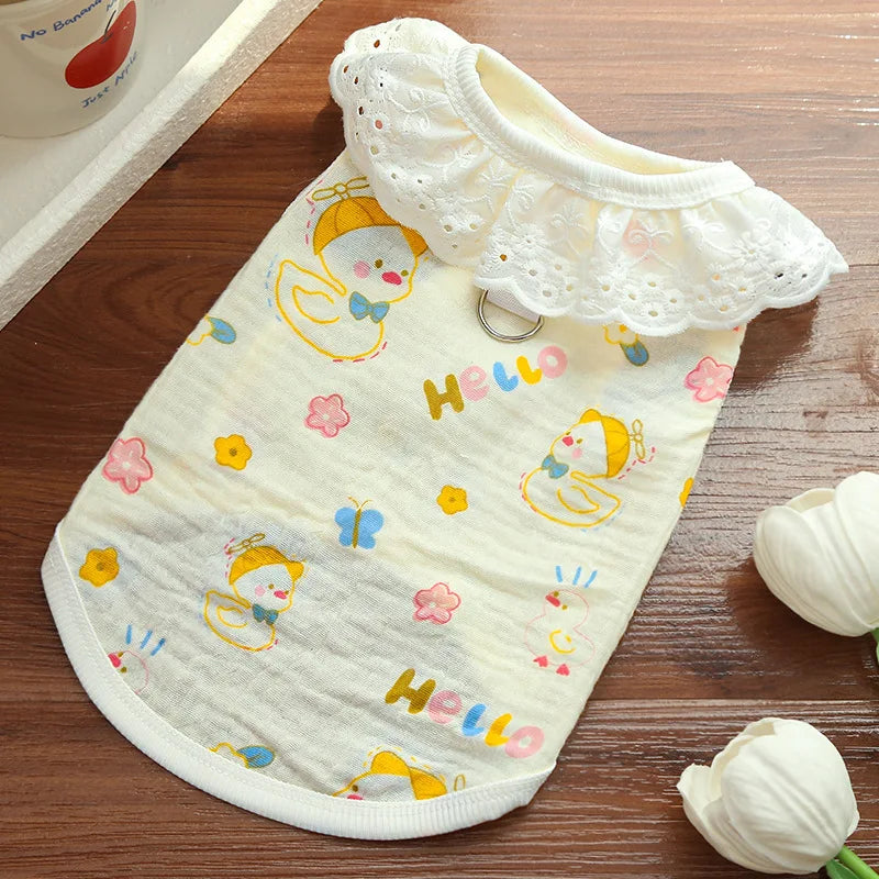 A cream-colored cotton dog vest with a white lace collar and yellow cartoon print, lying on a wooden surface.