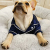 A light-colored dog is wearing a navy blue silk pet pajama shirt while lying on a fluffy white surface.