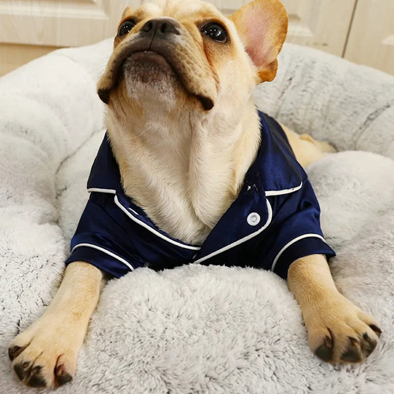 A light-colored dog is wearing a navy blue silk pet pajama shirt while lying on a fluffy white surface.