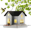A solar outdoor bird feeder lantern house with a dark roof and light gray siding is shown on a tray with green leaves overhea