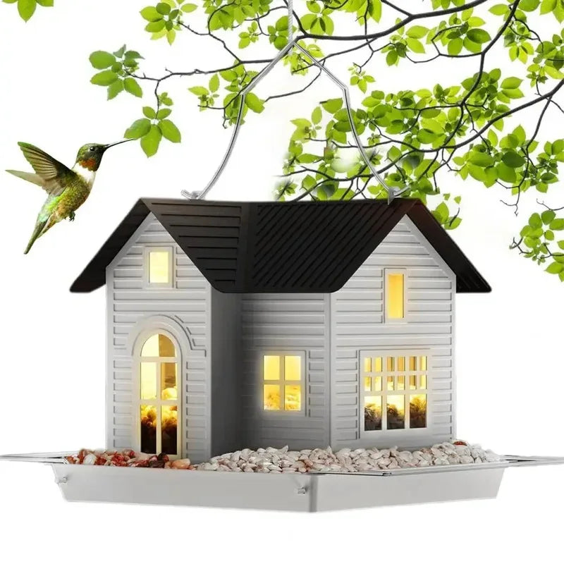 A solar outdoor bird feeder lantern house with a dark roof and light gray siding is shown on a tray with green leaves overhea