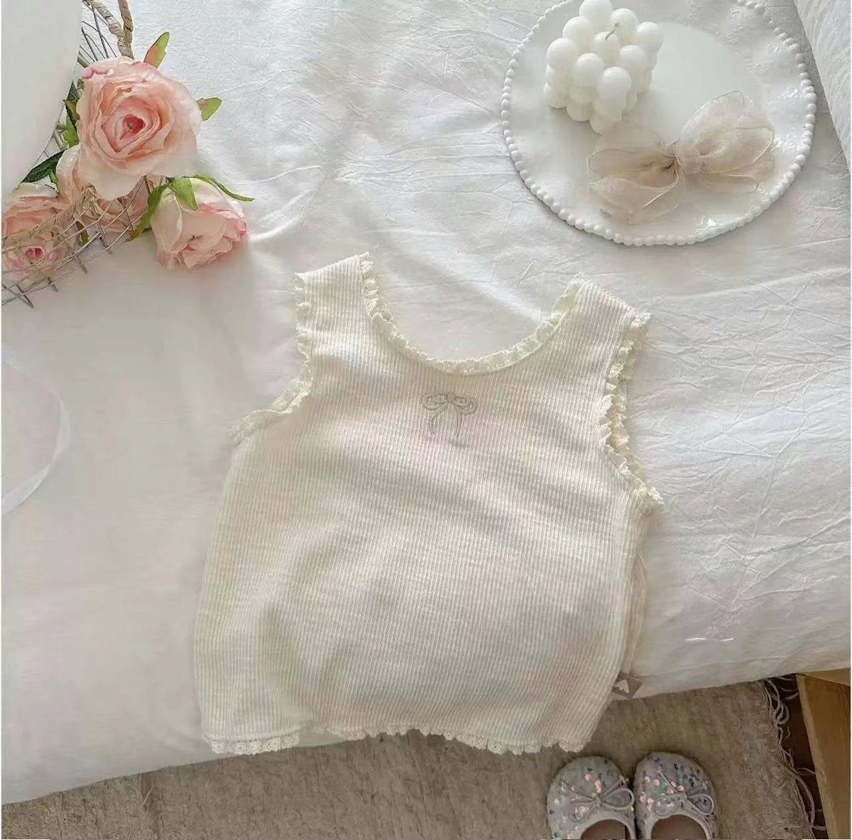 A white knitted mini vest for a baby girl with lace trim, displayed on a white surface next to pink flowers and a bow.