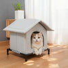 A gray heated outdoor cat shelter with a pitched roof is placed on a wooden floor next to a window.