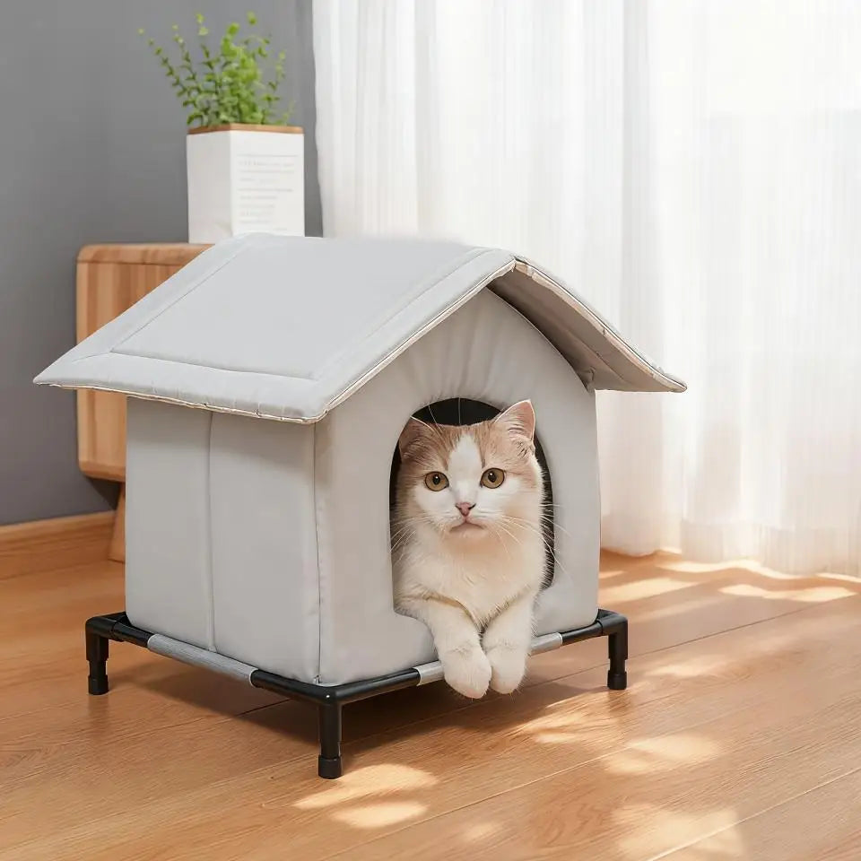 A gray heated outdoor cat shelter with a pitched roof is placed on a wooden floor next to a window.