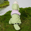 Un cachorro de perro con impermeable verde claro, collar rosa y capucha, tumbado sobre césped verde.