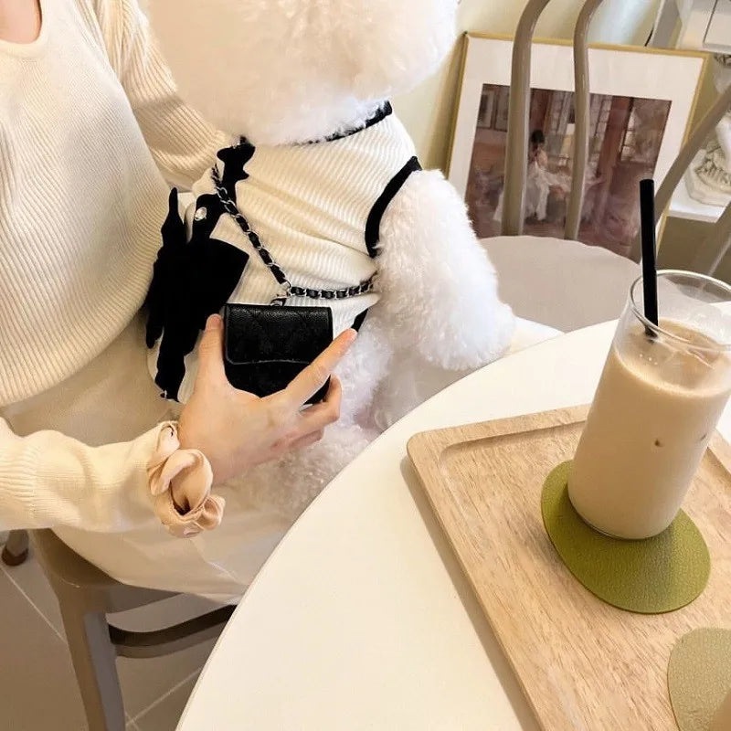 A person is holding a white fluffy dog dress with a black lace bow and a chain strap, next to a glass of iced beverage.