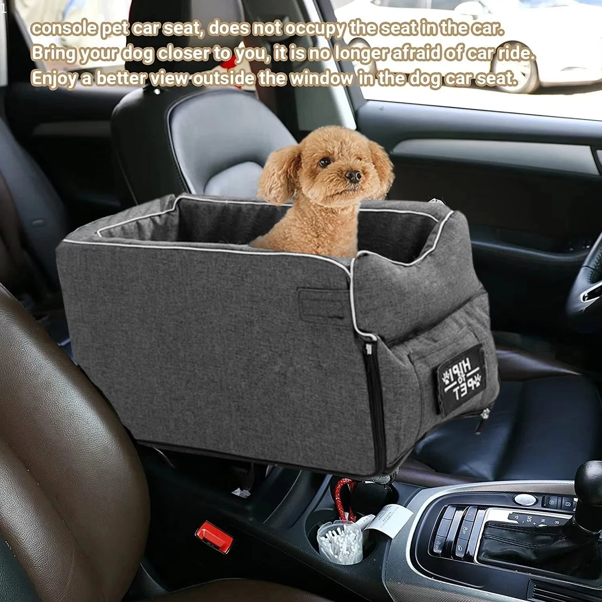 A small, light brown dog is sitting in a gray portable pet car seat placed in the back seat of a car.