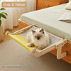 A fluffy light-colored cat is relaxing on a solid wood hanging cat hammock with yellow fabric in a bright room.