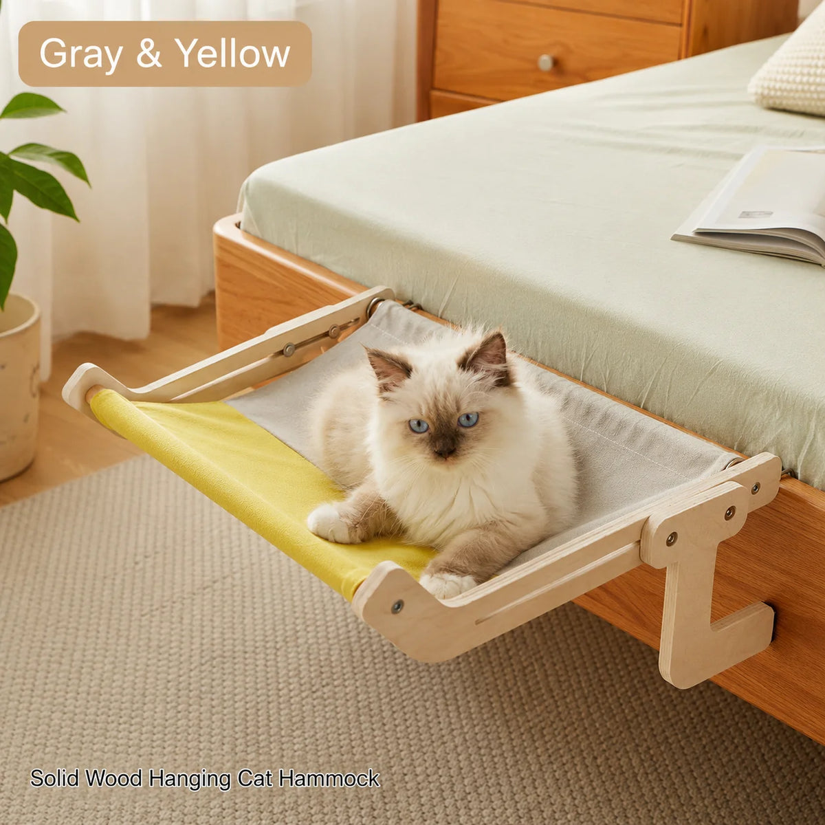 A fluffy light-colored cat is relaxing on a solid wood hanging cat hammock with yellow fabric in a bright room.