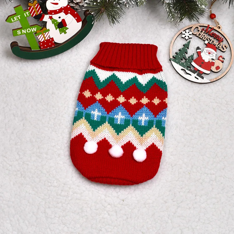 A knitted red dog sweater featuring geometric patterns and white pom-poms, displayed next to Christmas ornaments.