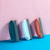 Three reusable pet hair remover brushes in dark teal, light blue, and terracotta against a pink background.