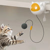 A small orange and white robotic cat toy is suspended, with a black string leading to a gray ball, near a tabby kitten.