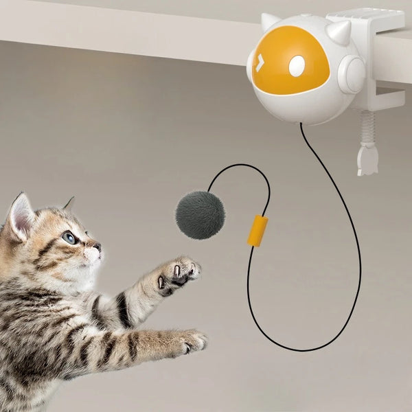 A small orange and white robotic cat toy is suspended, with a black string leading to a gray ball, near a tabby kitten.