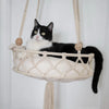 A black and white cat is resting in a macrame hammock pet bed suspended from a white wall.
