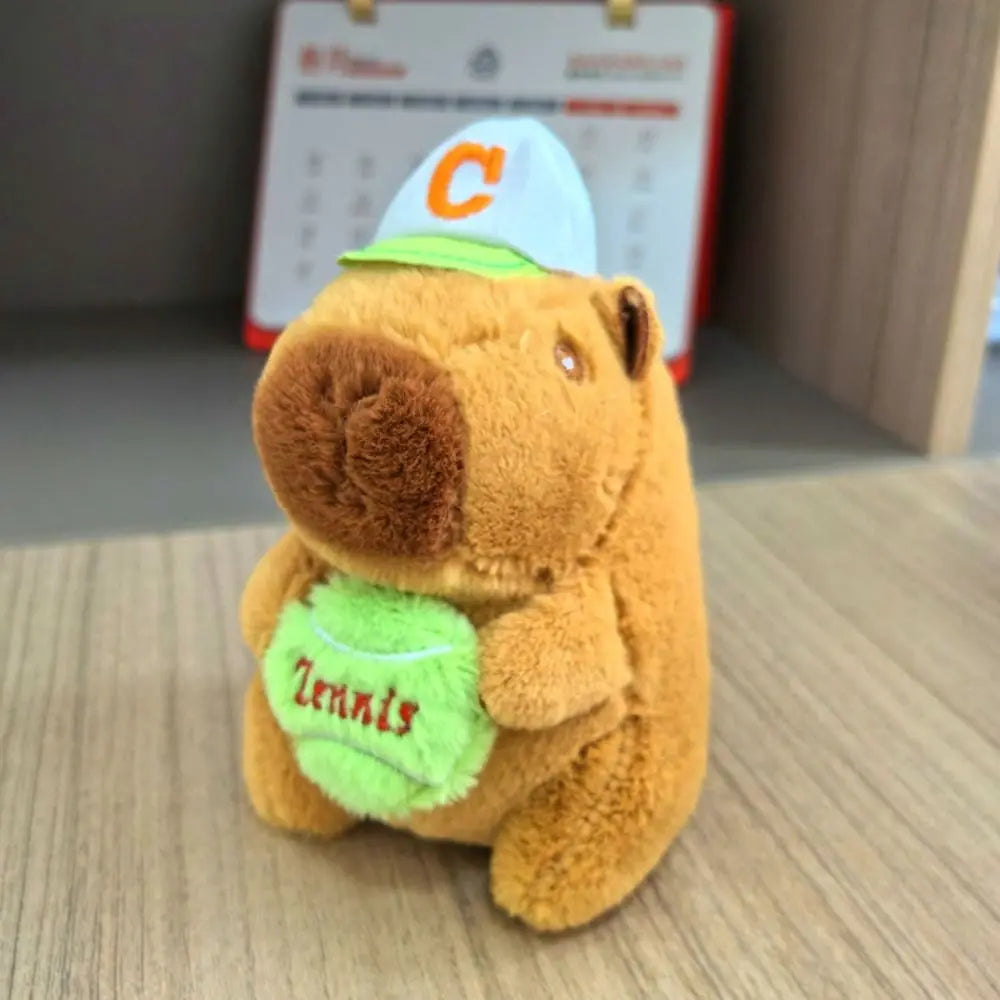 An orange capybara plush doll wearing a green hoodie and a white baseball cap with an orange letter C.