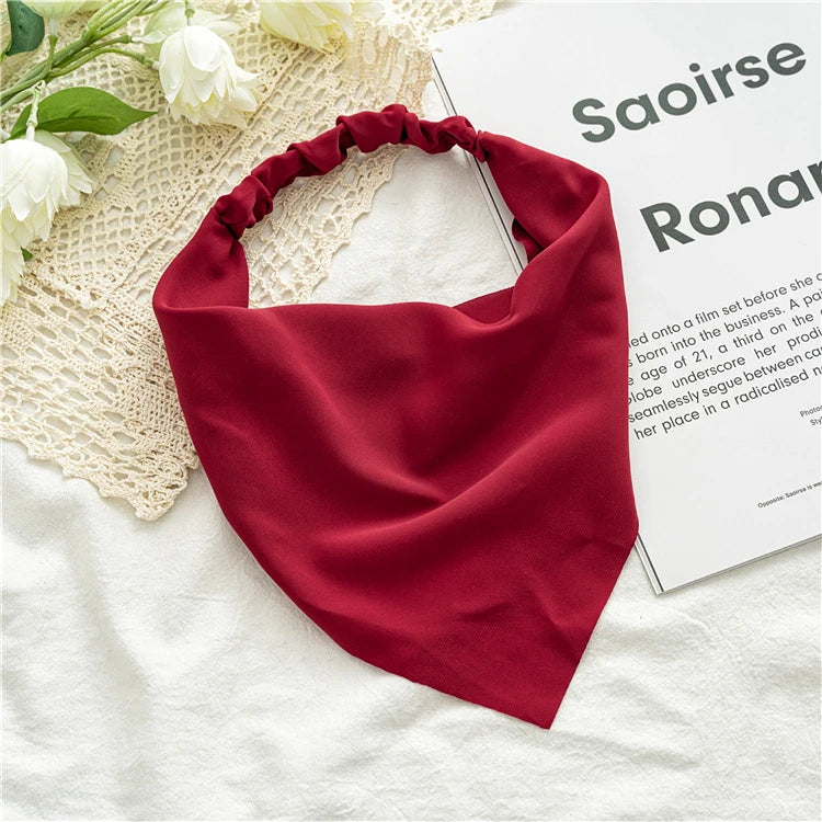 A deep red bohemian paisley triangle head scarf laid on a white fabric next to a piece of paper.