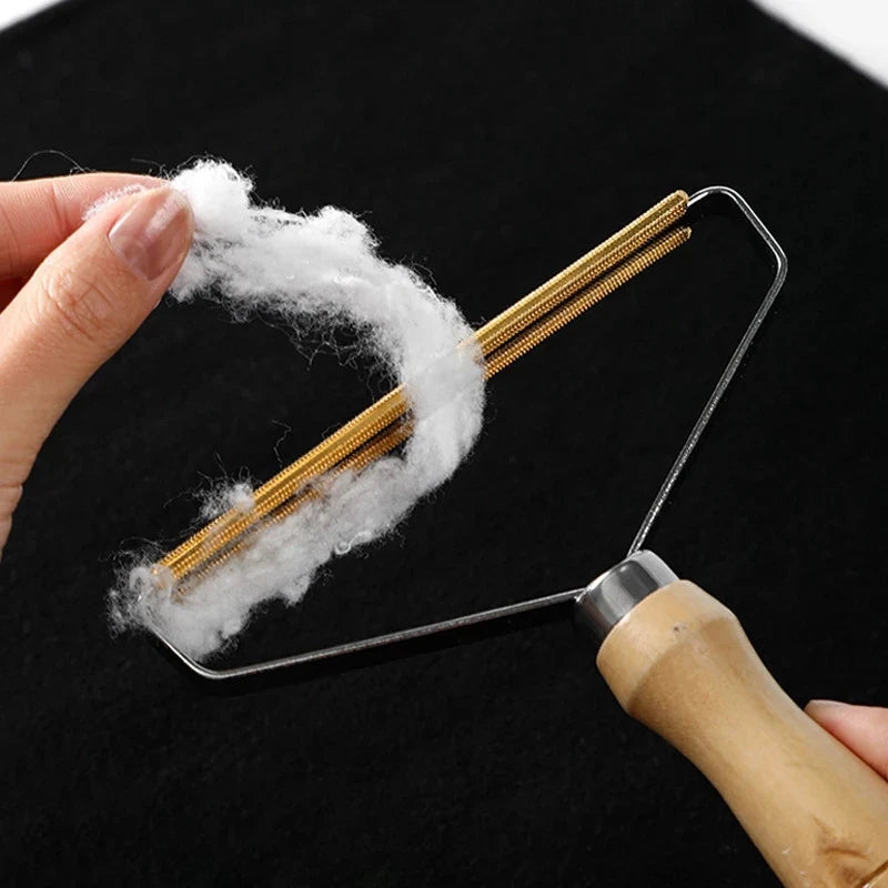 A hand holds a metal lint remover brush with gold-colored bristles, removing white lint from fabric.
