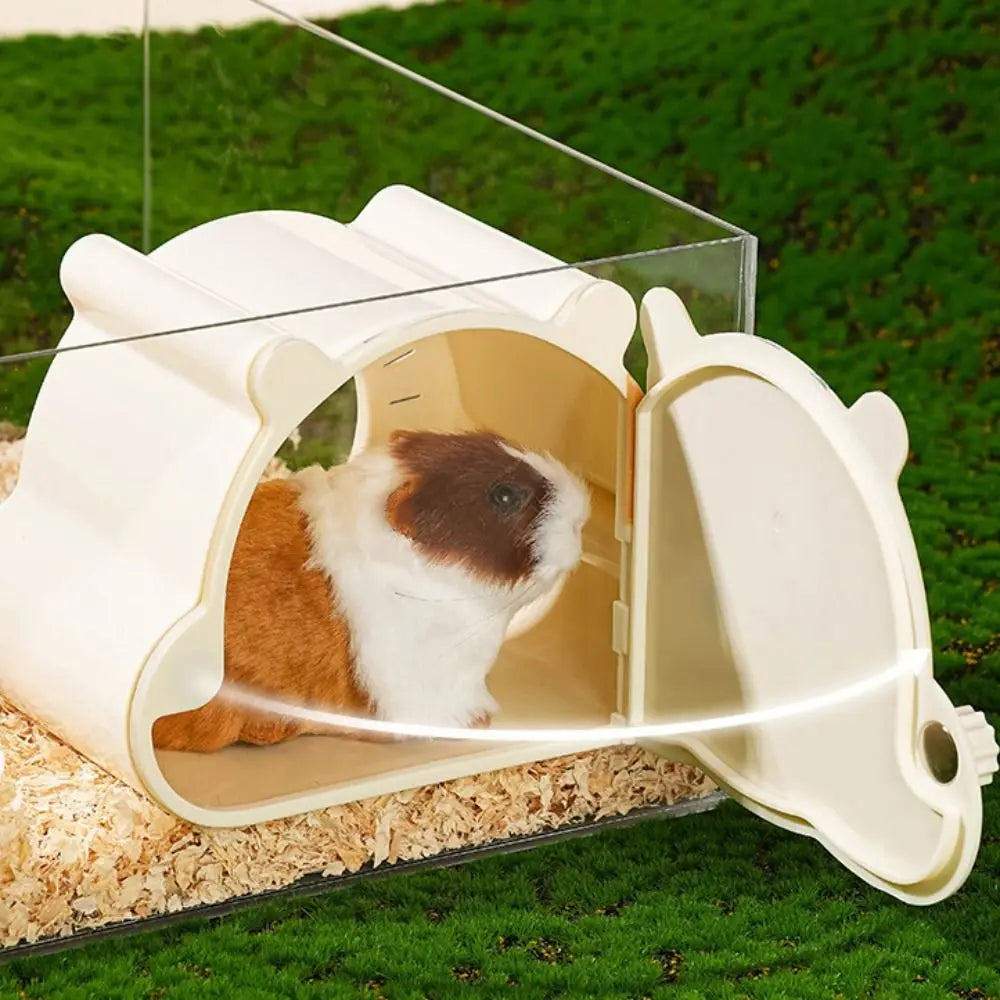 A white cartoon hamster hideout house with a viewing window is shown resting on green grass.