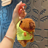 A hand holds a brown capybara plush doll wearing a lime green hoodie and gold hoop earrings against a patterned background.