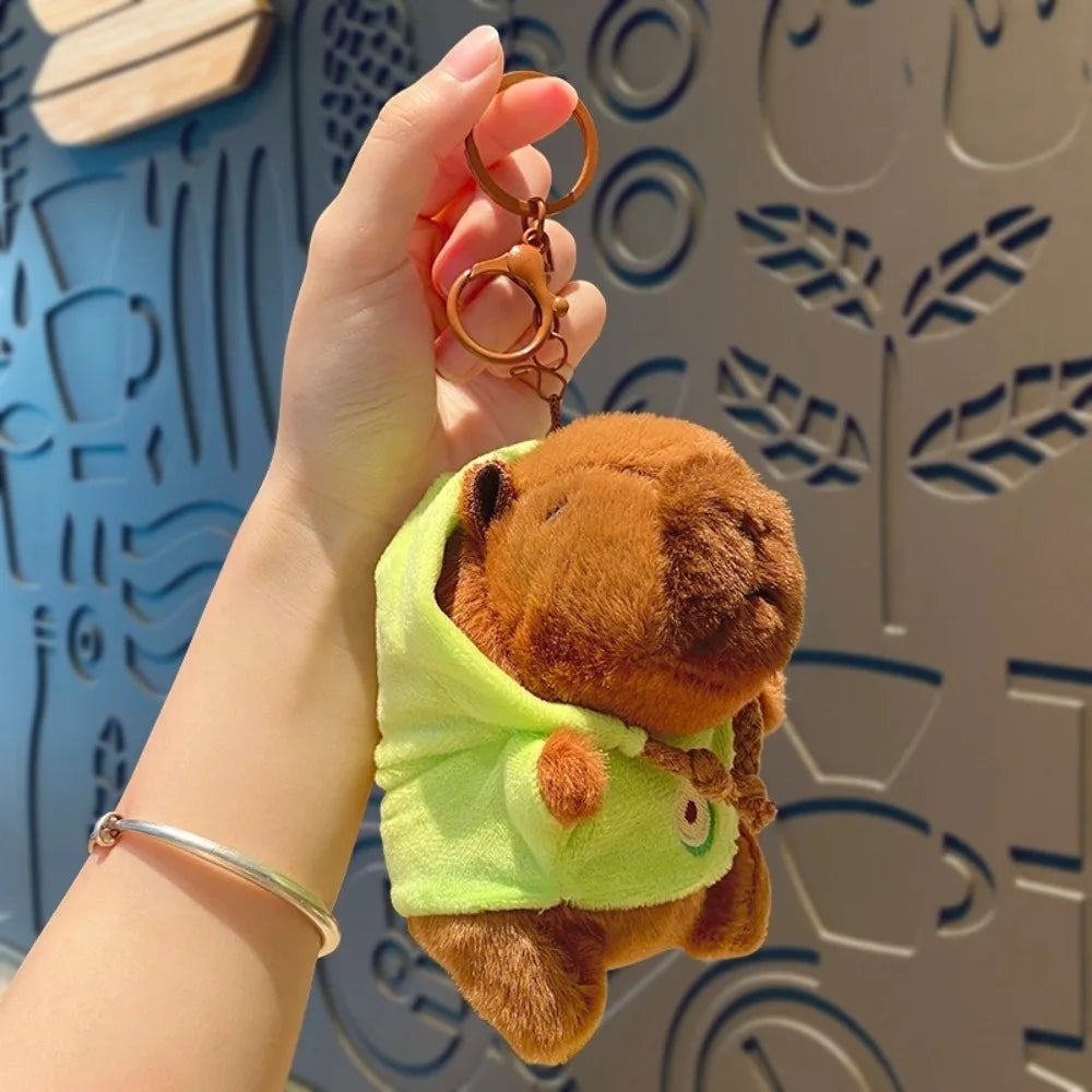 A hand holds a brown capybara plush doll wearing a lime green hoodie and gold hoop earrings against a patterned background.