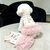 Ein weißes flauschiges Hundekleid mit pinkem Zierrand und Rüschen auf einer hellen Oberfläche.