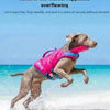 A dog wearing a pink and blue life jacket is leaping in clear ocean water on a sandy beach.