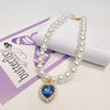 A delicate pearl necklace with a heart pendant displayed on a purple card.