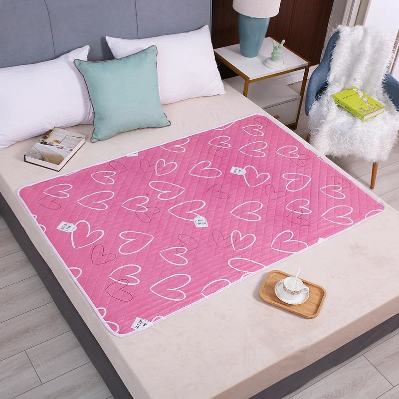A pink heart-patterned waterproof bed protector pad is displayed on a light-colored table with pillows and a white fluffy bla