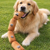 A golden retriever is holding a brown plush sausage dog chew toy with smiling faces on it in green grass.