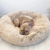 A fawn-colored French Bulldog is resting inside a fluffy, round pet bed.