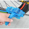 A hand holds a blue bike chain cleaner with a 3-sided brush system, positioned near a bicycle wheel.