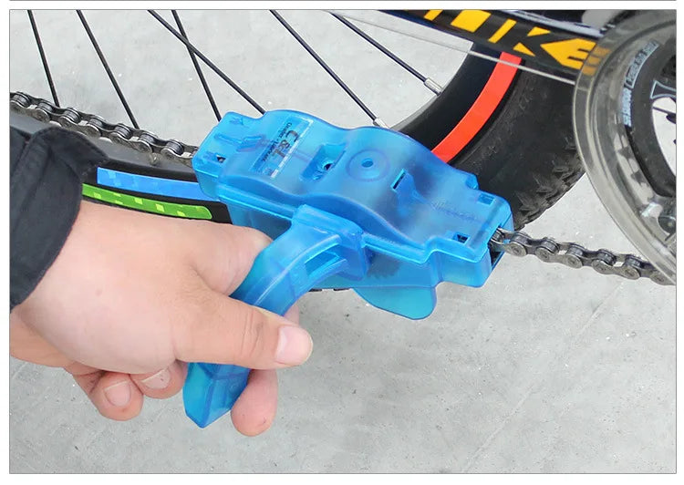 A hand holds a blue bike chain cleaner with a 3-sided brush system, positioned near a bicycle wheel.