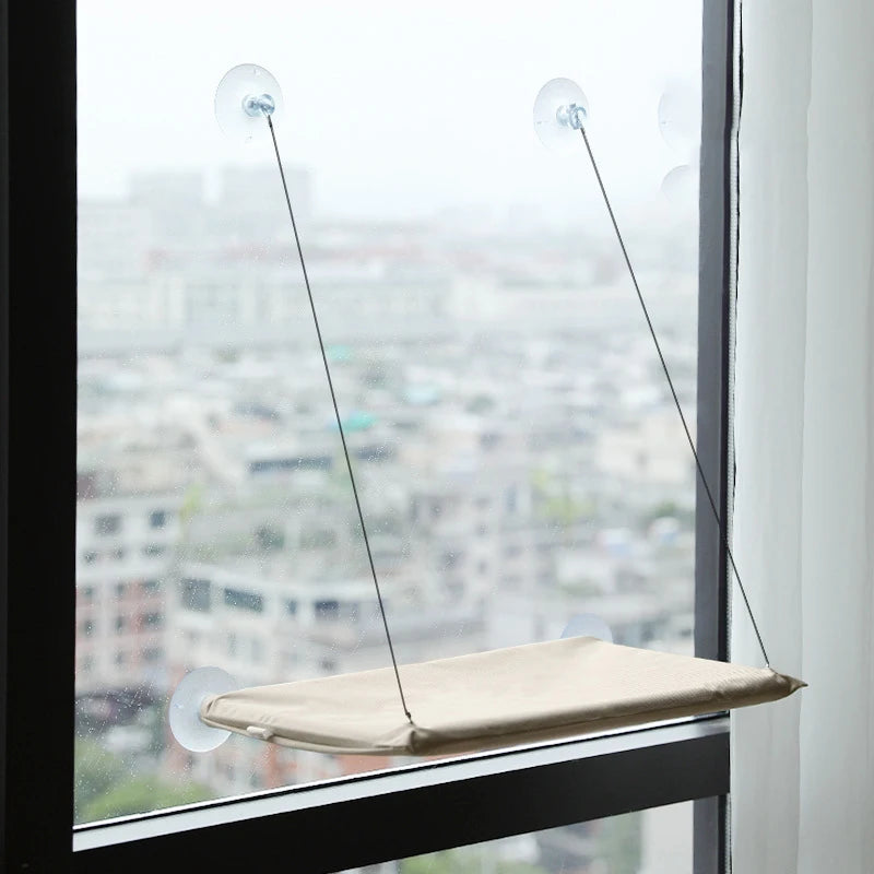 A beige hanging cat hammock suspended by thin wires from suction cups in a window.