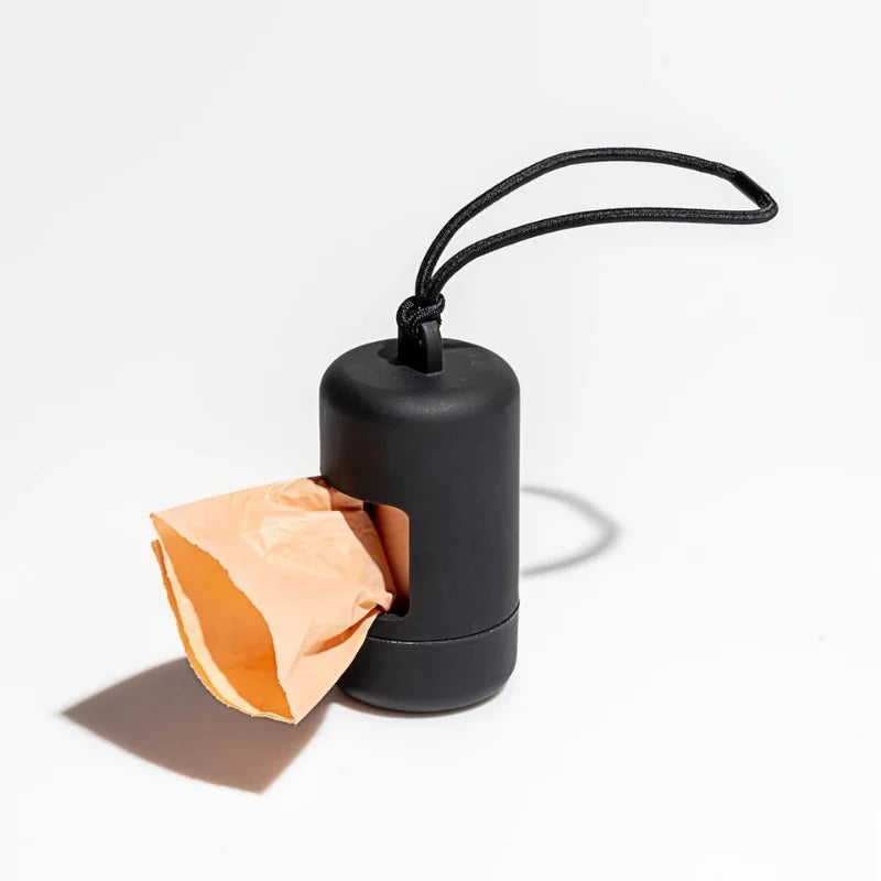 A black portable dog poop bag dispenser with a thin black loop attached, next to a crumpled piece of orange paper.