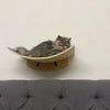 A fluffy gray cat is resting inside a wooden modular wall shelf designed with a bridge and hammock.