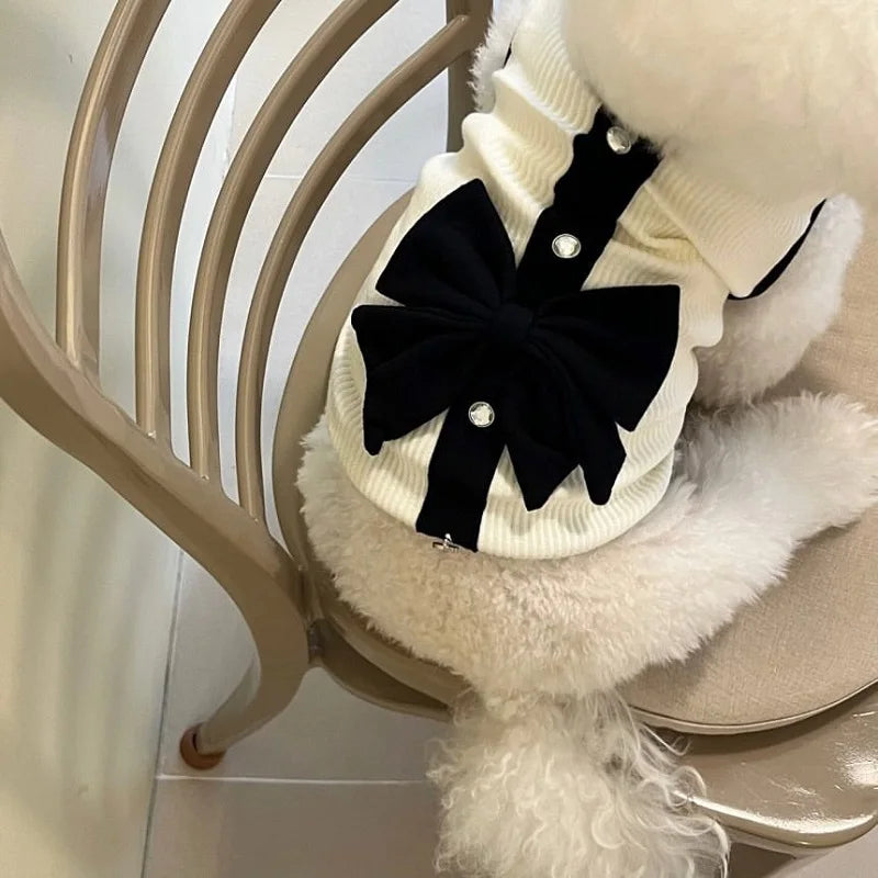 A white dog dress featuring a black lace bow and fluffy white trim, displayed on a wooden chair.