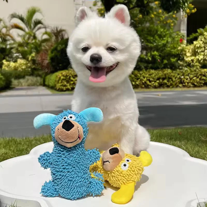 A white fluffy dog sits next to two blue and yellow plush toys on a white surface.