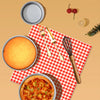 A round aluminum cake pan set with a baked cake, utensils, and a whisk displayed on a red and white checkered napkin.