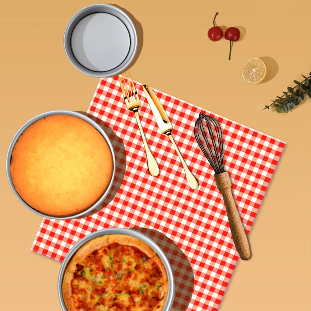 A round aluminum cake pan set with a baked cake, utensils, and a whisk displayed on a red and white checkered napkin.