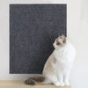 A white and gray cat is sitting in front of a dark gray textured scratching mat.