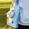 A white cat is visible inside a light blue portable crossbody pet carrier bag being carried by a person outdoors.