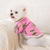 A white dog wearing a pink knit sweater with a brown bear pattern, resting on a cream blanket.