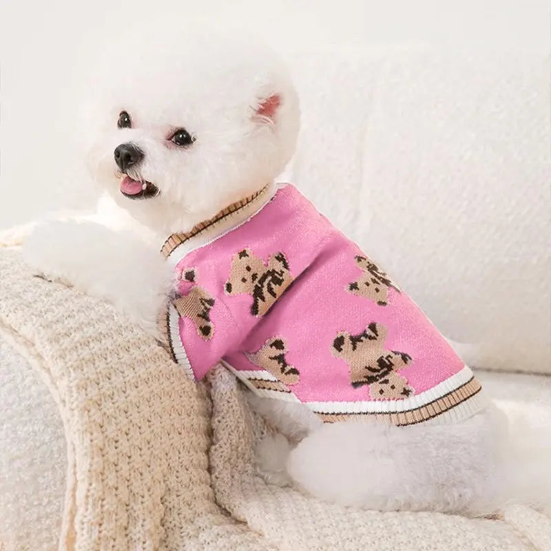 A white dog wearing a pink knit sweater with a brown bear pattern, resting on a cream blanket.
