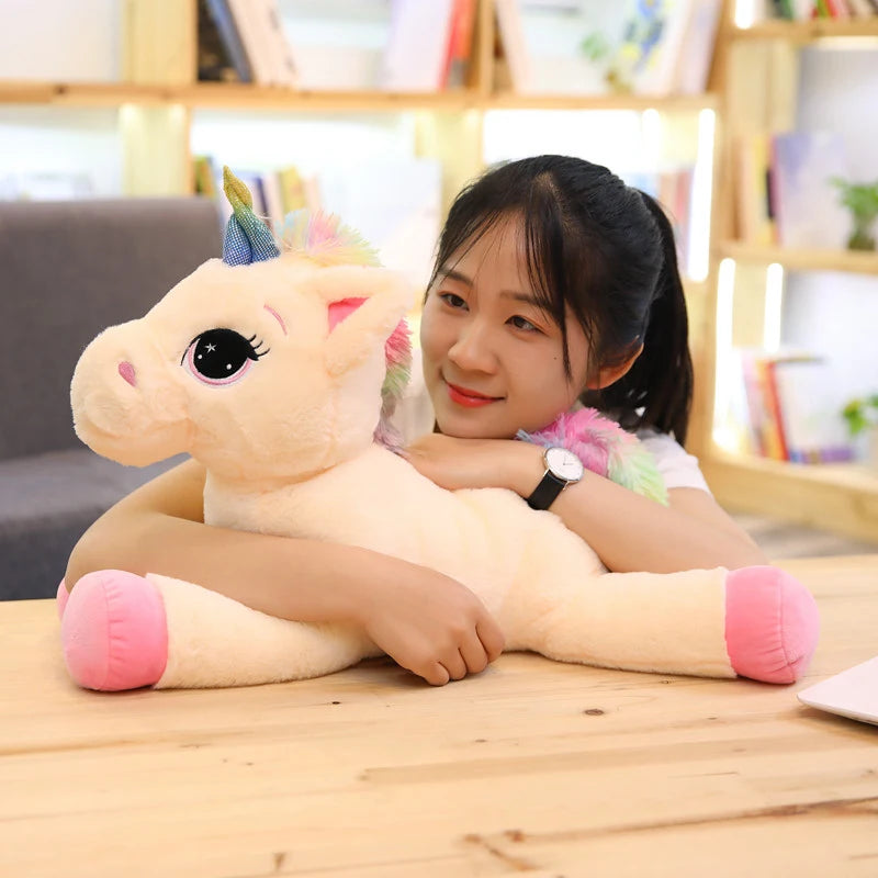 A young woman is cuddling a light pink unicorn plush toy with a rainbow mane and horn on a wooden table.