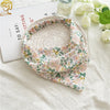 A floral patterned triangle head scarf is displayed on a white cloth next to a card and some white roses.
