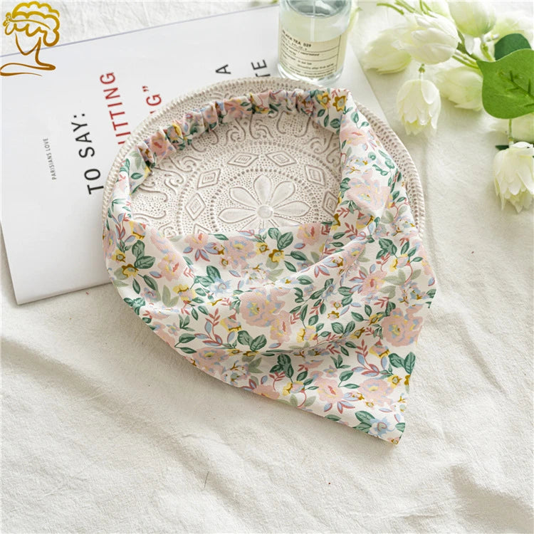 A floral patterned triangle head scarf is displayed on a white cloth next to a card and some white roses.