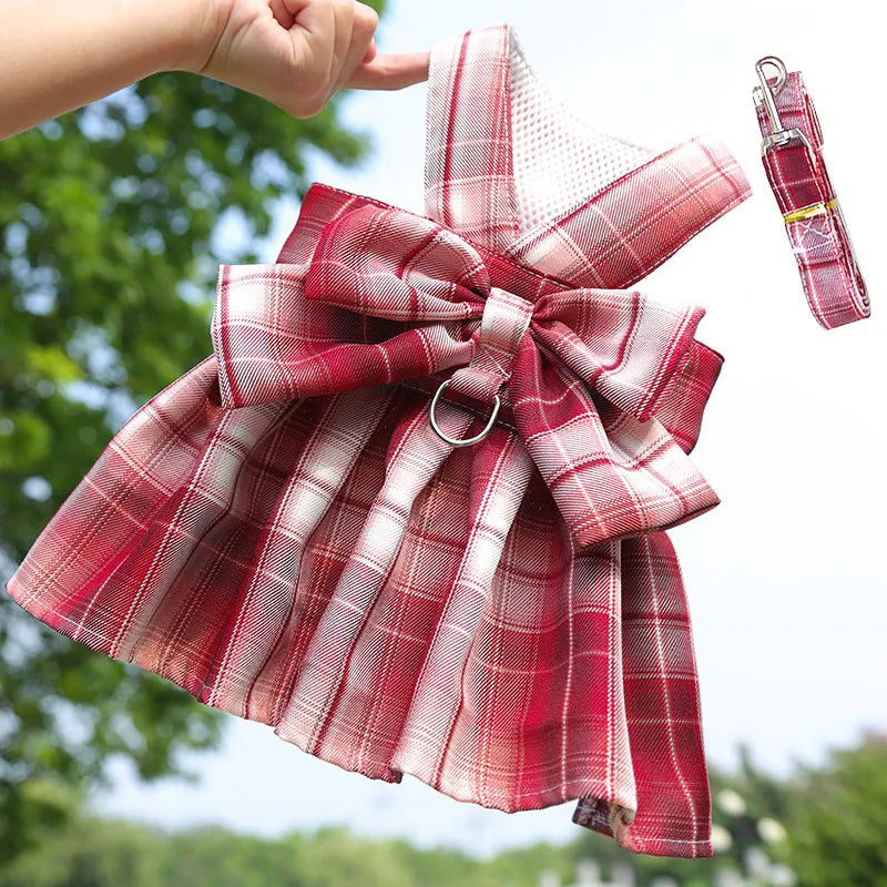 A red plaid pet dress-up outfit featuring a bow and a matching harness is shown against a green outdoor background.