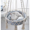 A fluffy white cat is resting inside a bohemian macrame hammock pet bed.