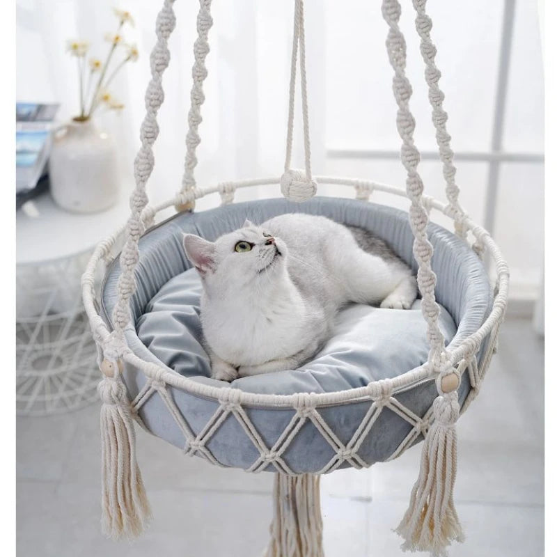 A fluffy white cat is resting inside a bohemian macrame hammock pet bed.