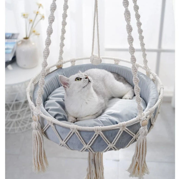 A fluffy white cat is resting inside a bohemian macrame hammock pet bed.